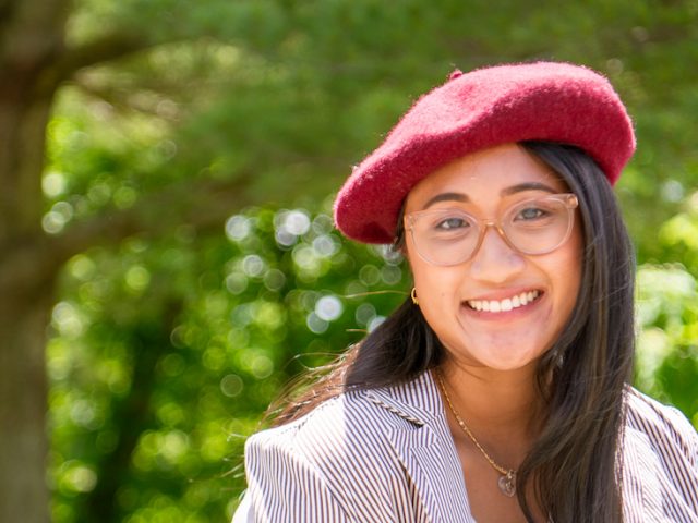 Stephanie wears glasses and a red beret smiling at the camera with a green forest in the background.