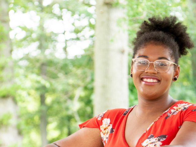 Jocelyn sits and smiles in front of a wooded area of campus.