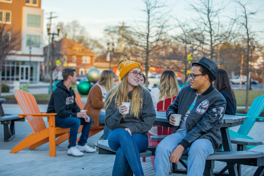 Two groups of students sit on chairs at Rowan Boulevard, bundled up for the weather, holding coffee cups.