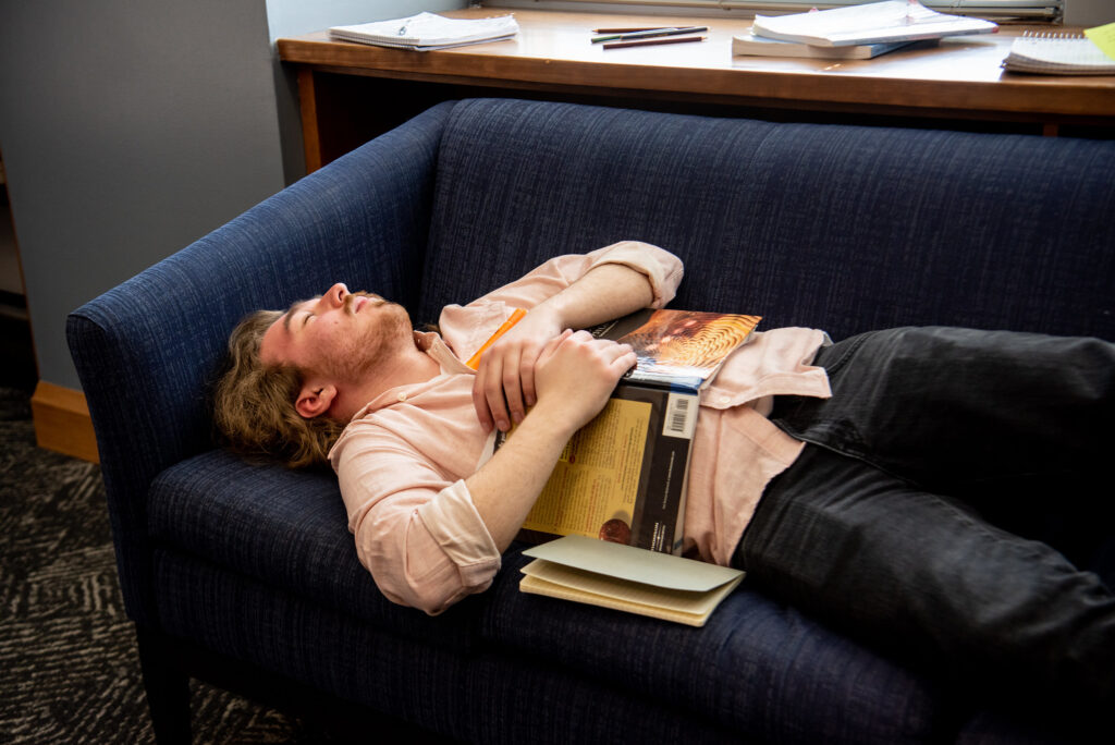 A student comedian from the improv club pretends to nap on a couch in the library with a book on his chest. The location is truly a favorite sleeping place for many students.