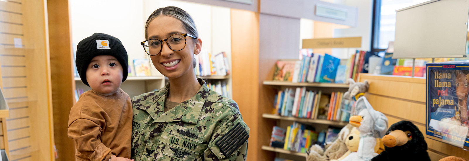 Rowan University student and mother holds her infant on her hip in the school bookstore, while smiling at the camera. She is wearing her Army reservist uniform.