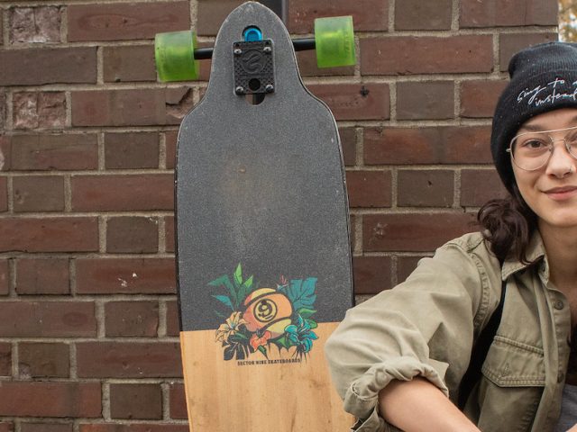 Maria sits with her longboard in front of a brick building on campus.