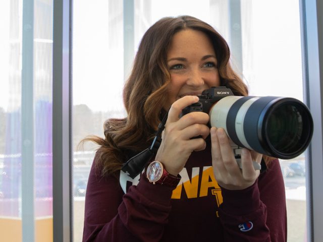 Ashley holds a DSLR camera with a long lens inside Business Hall.