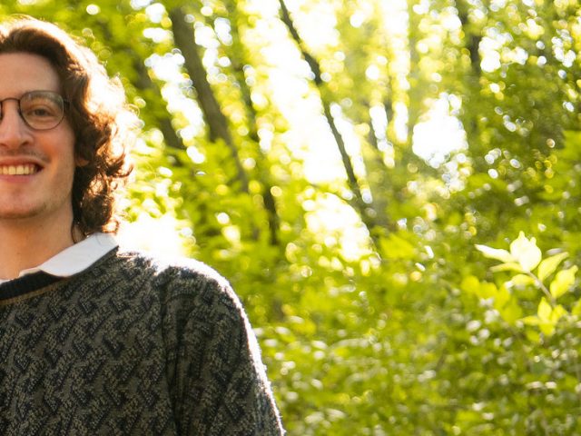 Brandon is smiling and gazing at the camera behind a forest.