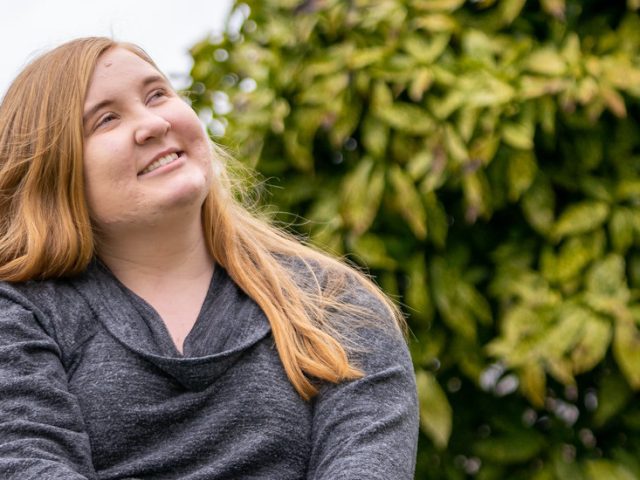 Rachel is smiling upwards and is in-between some shrubbery.