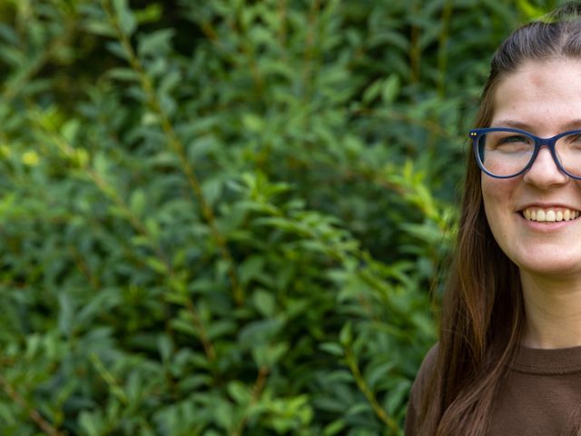 Sarah smiles with green shrubs in the background.