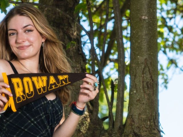 Skyla holds a Rowan University pennant against a wooded backdrop.