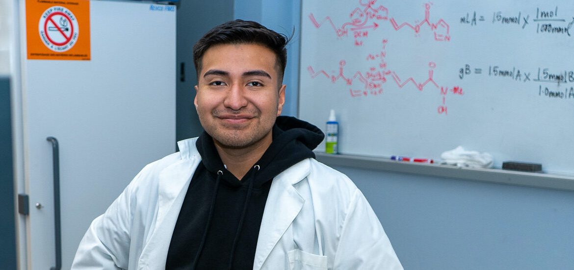 Daniel is sitting in a chemistry lab with his white lab coat and smiling directly at the camera.