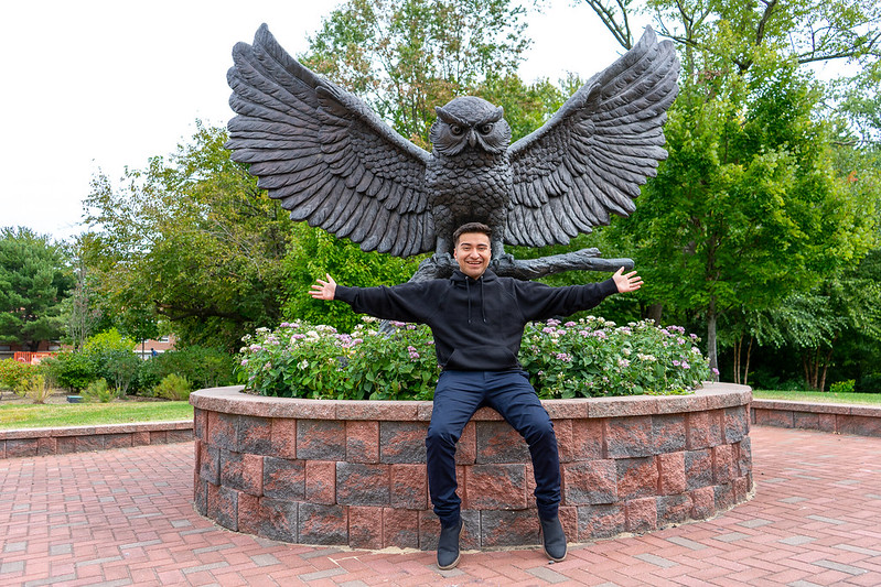 Daniel is sitting at the owl statue on campus and spreading his arms out like the sculpture.