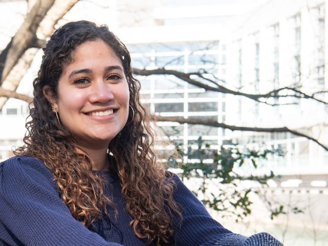 Maria is sitting and smiling with Rowan Hall in the background.