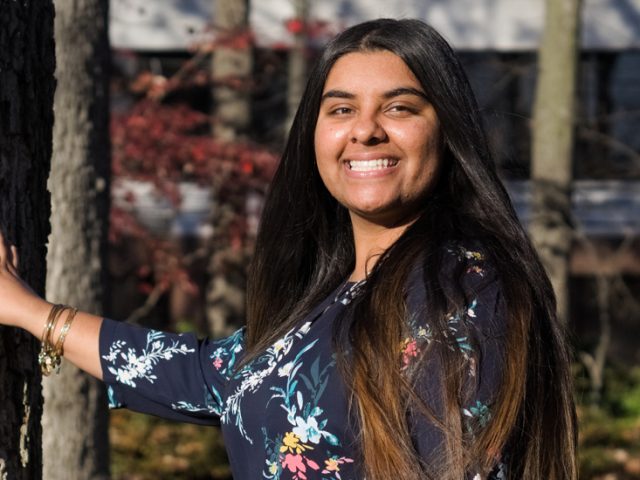 Riya stands beside a tree, smiling.