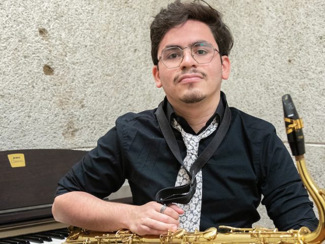 Rowan University student Jovan Rivera posing inside of Wilson Hall with a saxophone sitting in front of a piano.