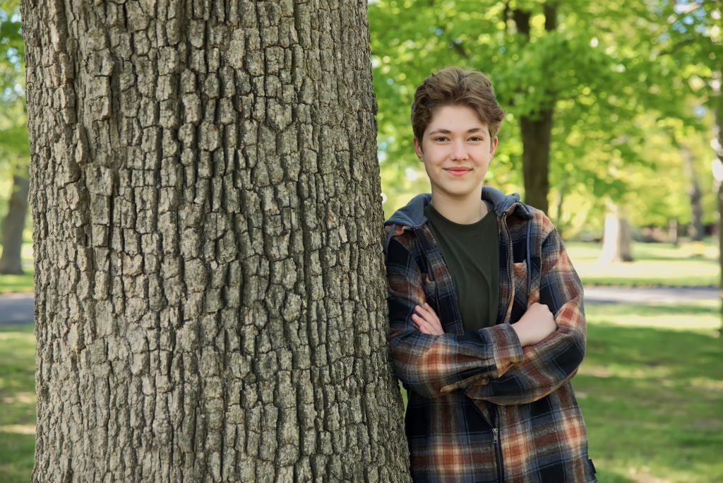 Kayden stands cross armed leaning against a tree.
