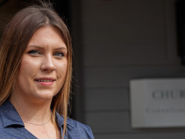 Kate stands in her work uniform in front of the sign for Churchill Construction Engineers