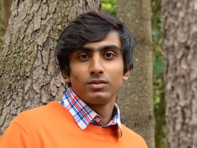 Kriish poses for an outdoor portrait in front of a tree while wearing a bright orange shirt.