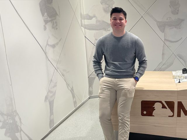 A student standing in front of a Major League Baseball work desk.