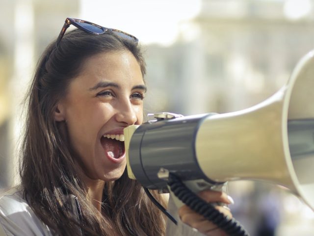 Stock image of a woman happily holding a megaphone to share a message.