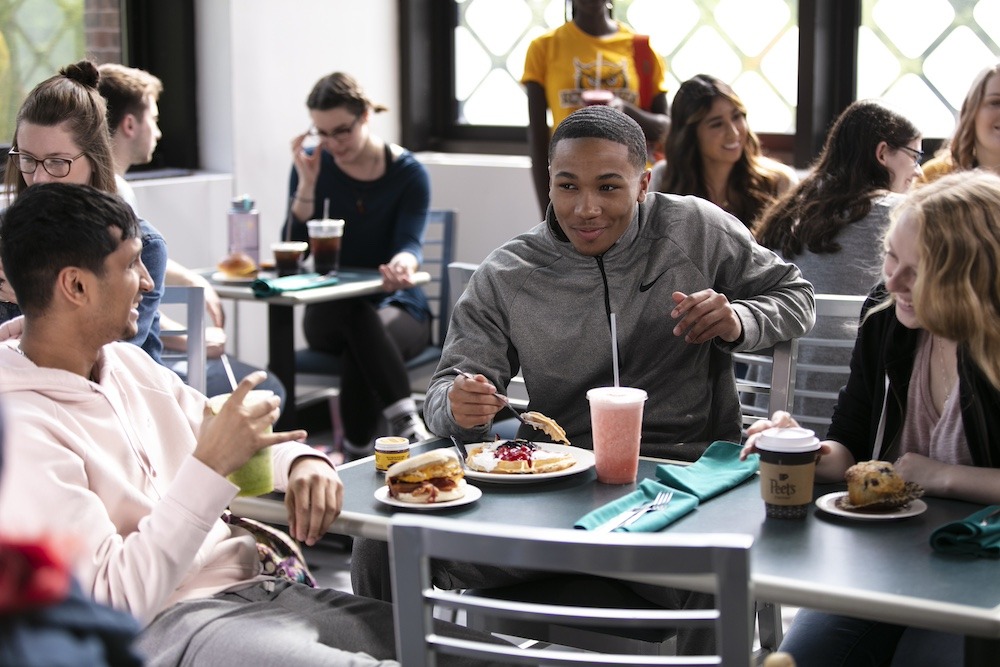 With a focus on three students eating and laughing over a meal at the Student Center, we see additional students at tables around them.