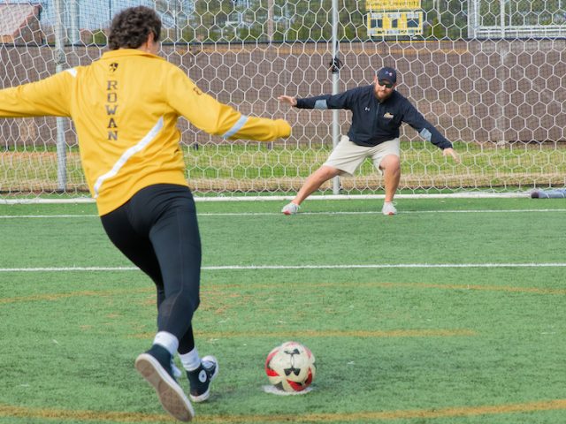 Miranda kicks a soccer ball into a soccer net as an alumnus, while wearing Rowan gold.