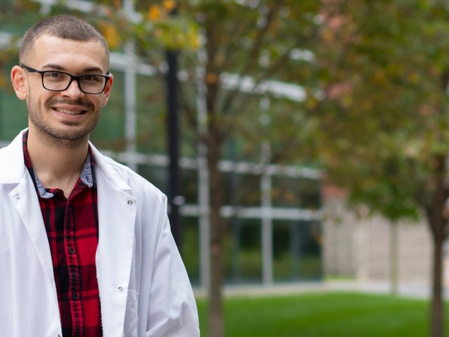 Nicholas stands outside the science building wearing a lab coat.