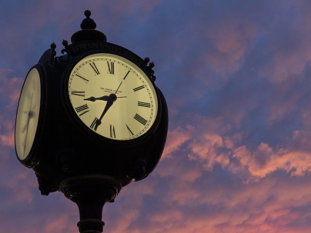 Close up of the face of a town clock with a dramatic sunset in the background.