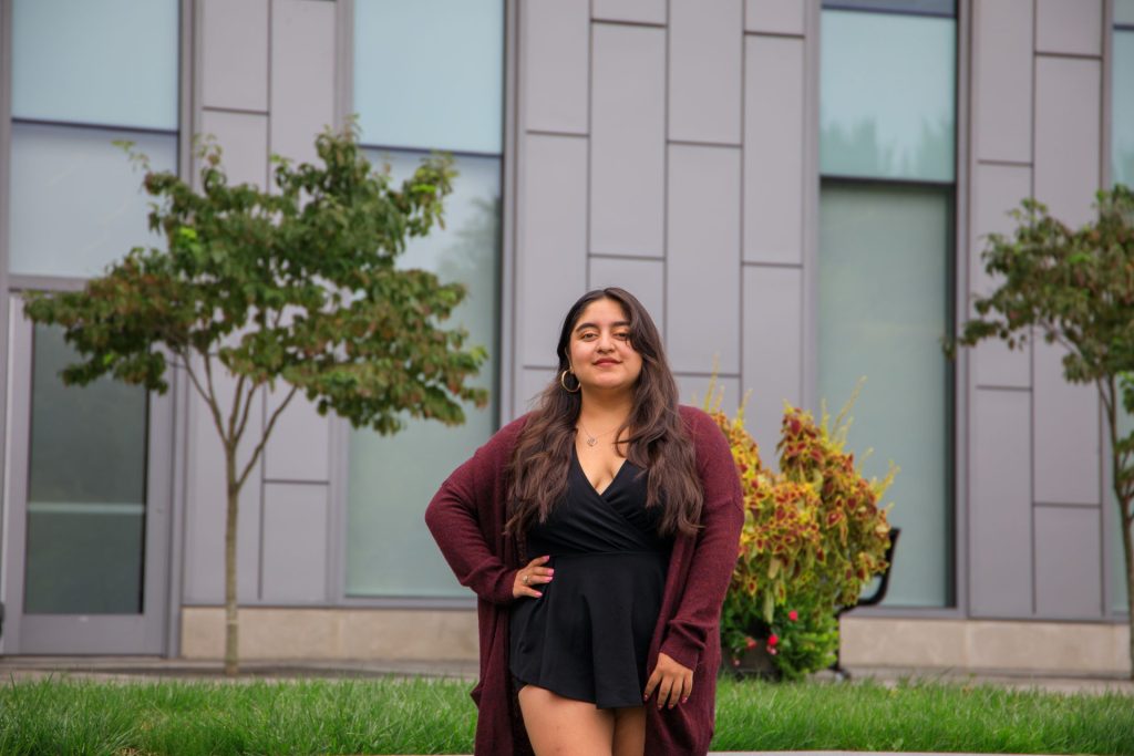 Law and justice major Domenica Gusqui Gavidia stands outside an academic building on the campus of Rowan University, wearing an oversized maroon cardigan and holding her hand on her hip. 