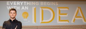 A student stands in front of a wall with the word "Idea" behind him, with his arms crossed across his chest.