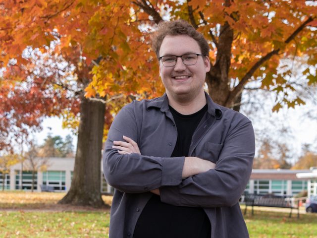 Alec stands arms crossed with vibrant trees behind.