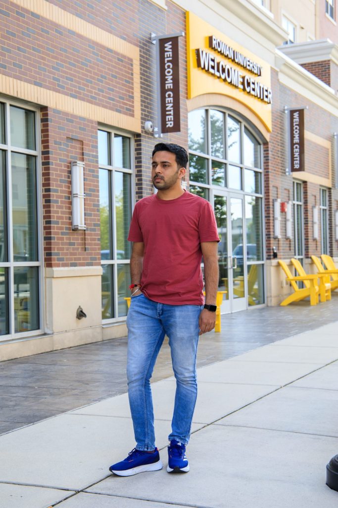 Dikeshkumar Kanani posing outside of the Welcome Center on Rowan Boulevard. He is seen in a red t-shirt with blue jeans.