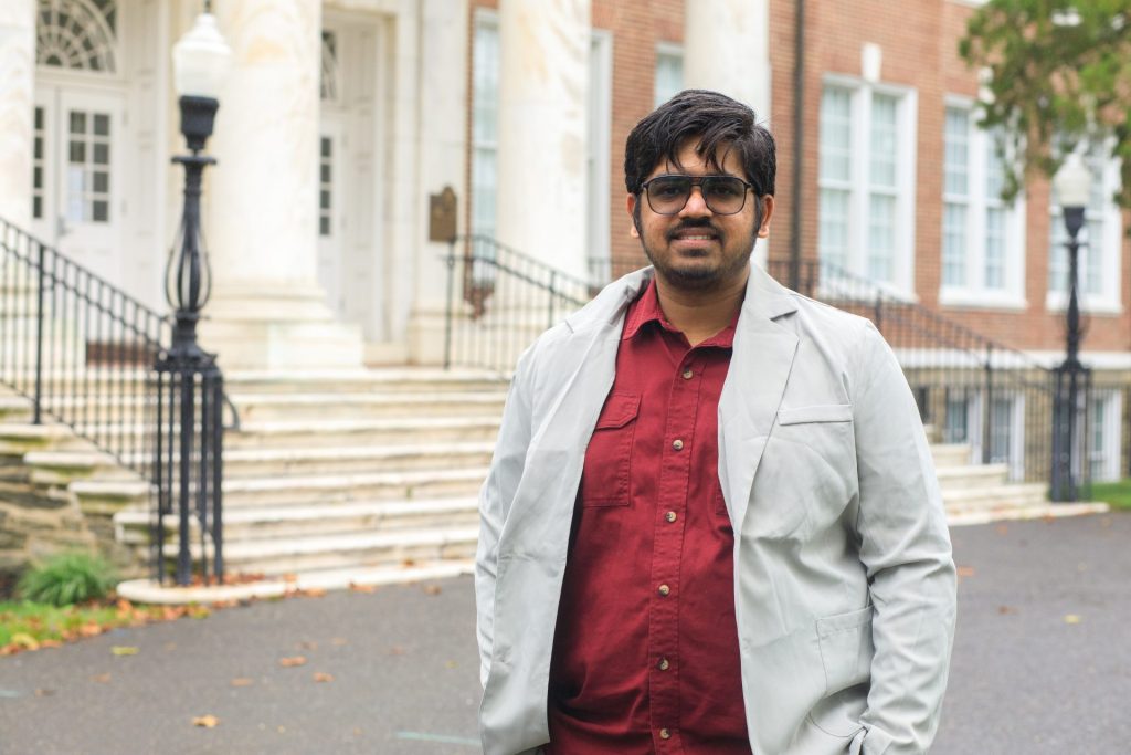 Guruprasanth standing outside of Bunce Hall in a red shirt and white coat jacket