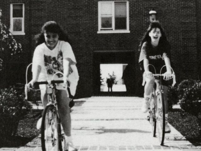 Black and white photo of two girls on bicycles outside a dorm.