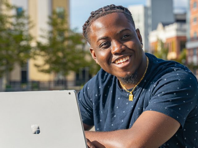 Samuel posing outside on Rowan Boulevard with his laptop and water bottle