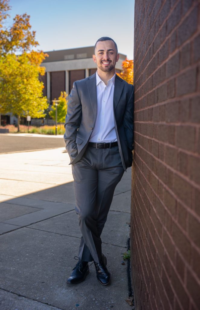 Shawn Ryan posing against a wall outside of Wilson Hall. He can be seen wearing a gray suit.
