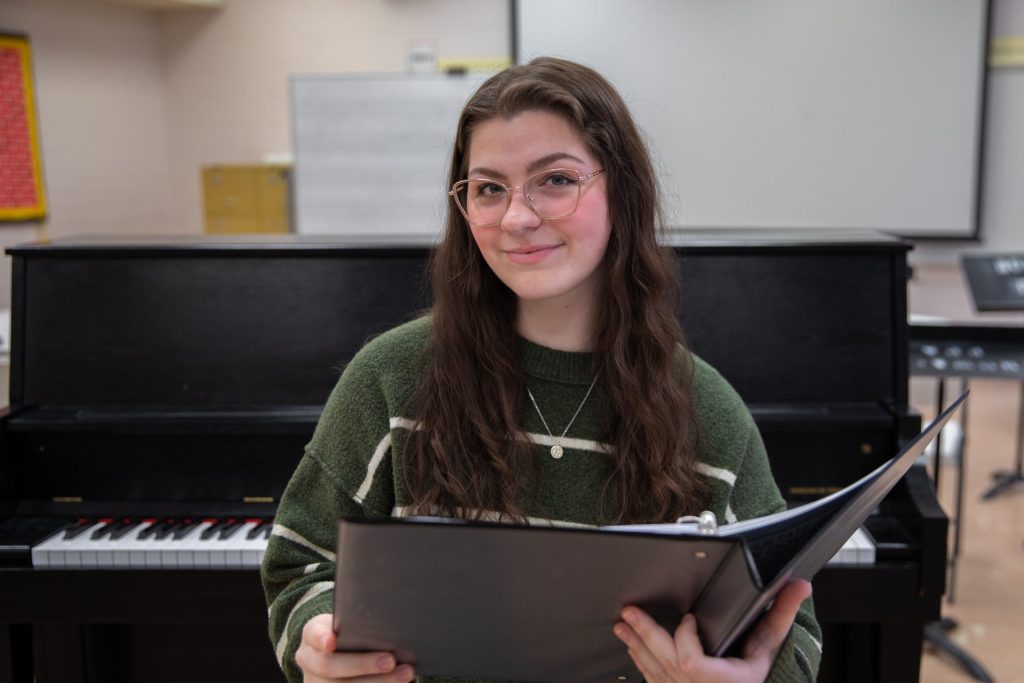 Hannah Potratz sitting down looking at the camera. She is holding her music binder. There is a piano behind her.