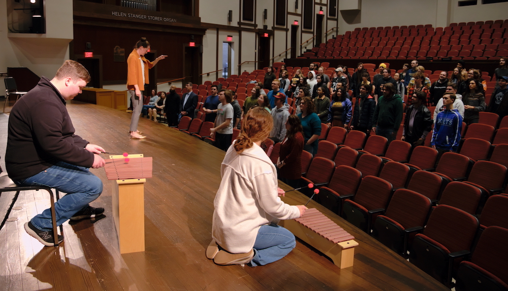 Two music class students on stage play instruments, while students in the audience seats stand and sing with direction from their professor. 