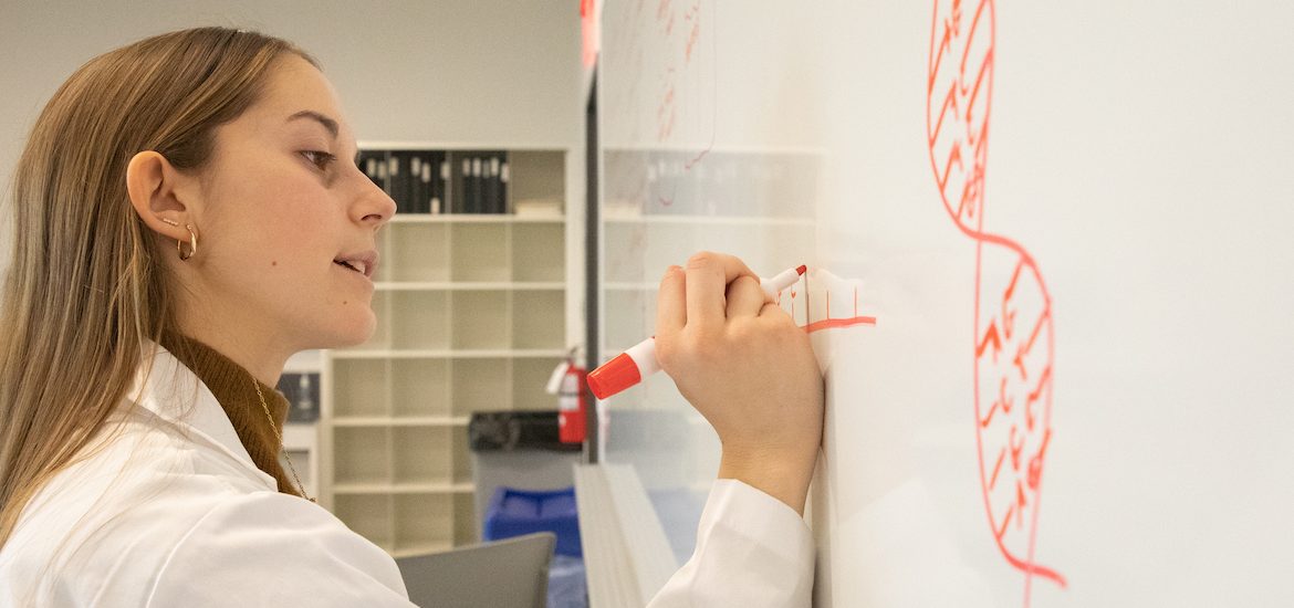 Mia drawing on the whiteboard. She is seen in a lab coat.