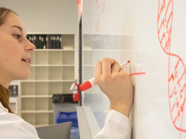 Mia drawing on the whiteboard. She is seen in a lab coat.