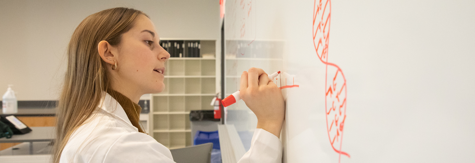 Mia drawing on the whiteboard. She is seen in a lab coat.