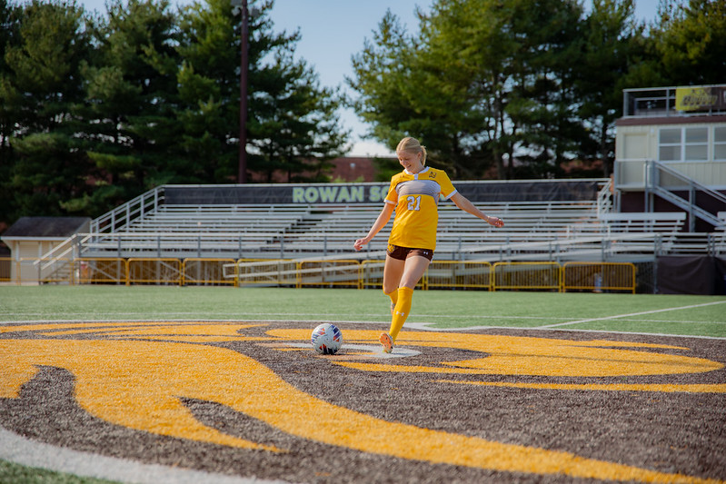 A student practices soccer on the soccer field, while wearing her Rowan yellow uniform. 