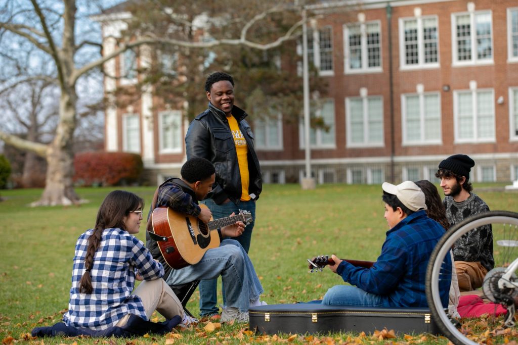 Six students sitting on Bunce Green. One is playing the guitar and the rest are surrounding the student.