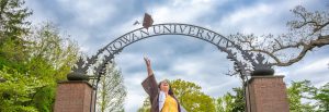 A student wearing a Rowan yellow dress and Rowan brown graduation gown tosses her graduation cap in the air in front of the Rowan University arch sign.