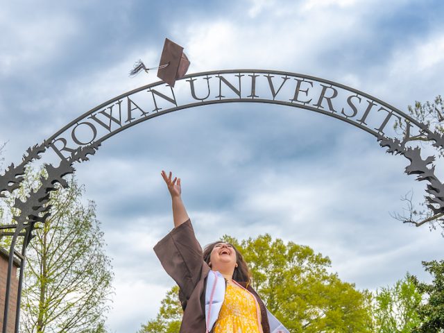 A student wearing a Rowan yellow dress and Rowan brown graduation gown tosses her graduation cap in the air in front of the Rowan University arch sign.