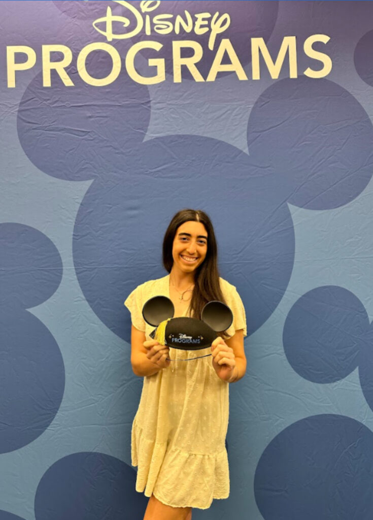 Sydney standing in front of a 'Disney Programs' sign, holding a pair of mouse ears.