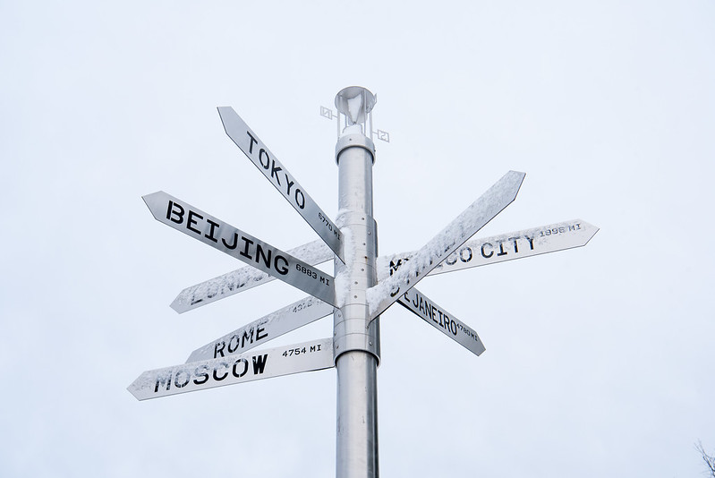 A silver directional road sign showing 7 different arrows and how many miles to international cities, such as Moscow 4,754 miles. 