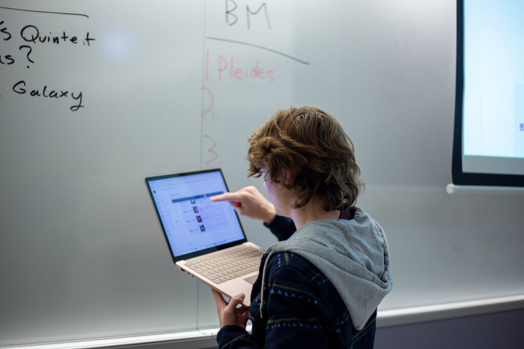 A person pointing at a laptop screen in front of a whiteboard.