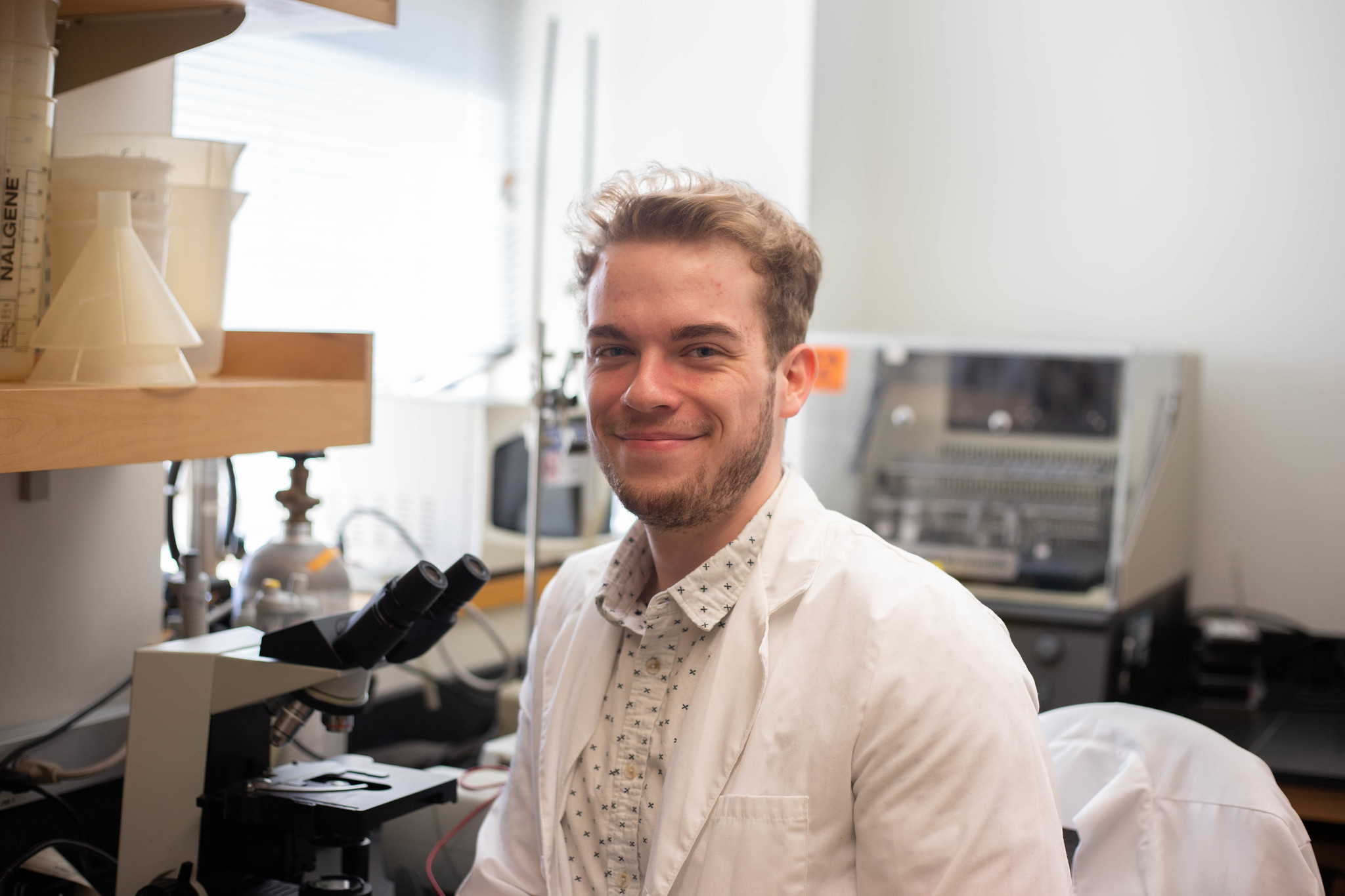 Rowan University biological sciences major Brian Libby sits in a lab, smiling at the camera, wearing a lab coat while seated. 