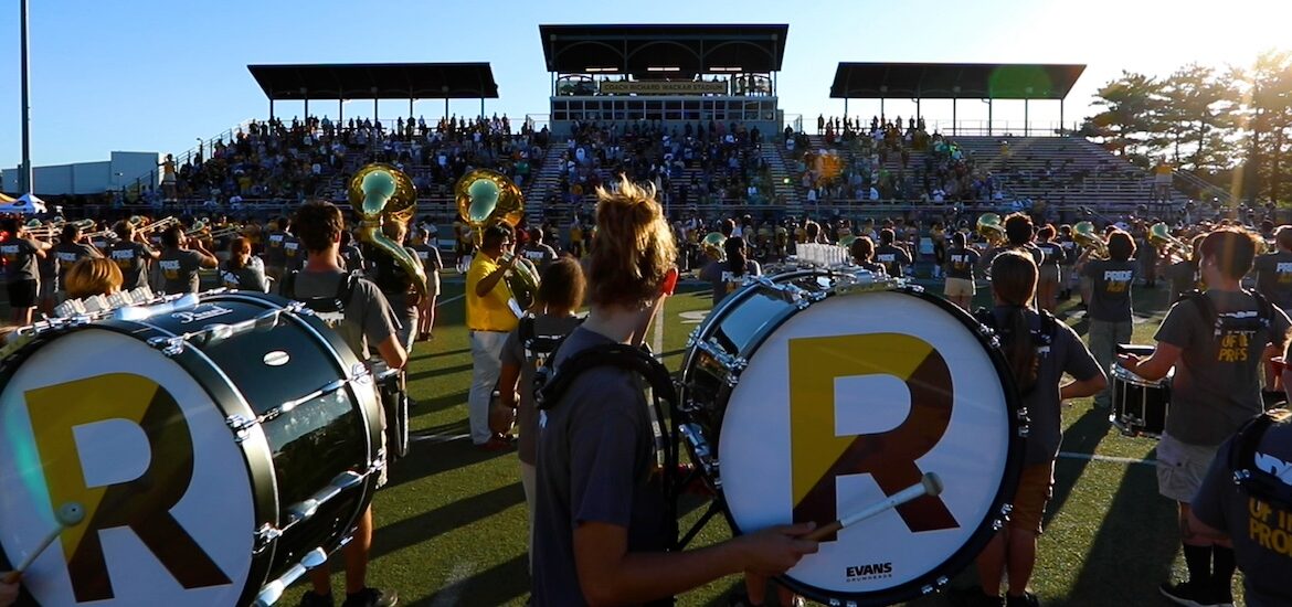 A side view of drum majors playing a drum during marching band, with a giant R on the side of the drum for Rowan.