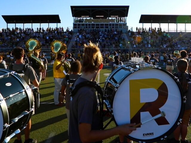 A side view of drum majors playing a drum during marching band, with a giant R on the side of the drum for Rowan.