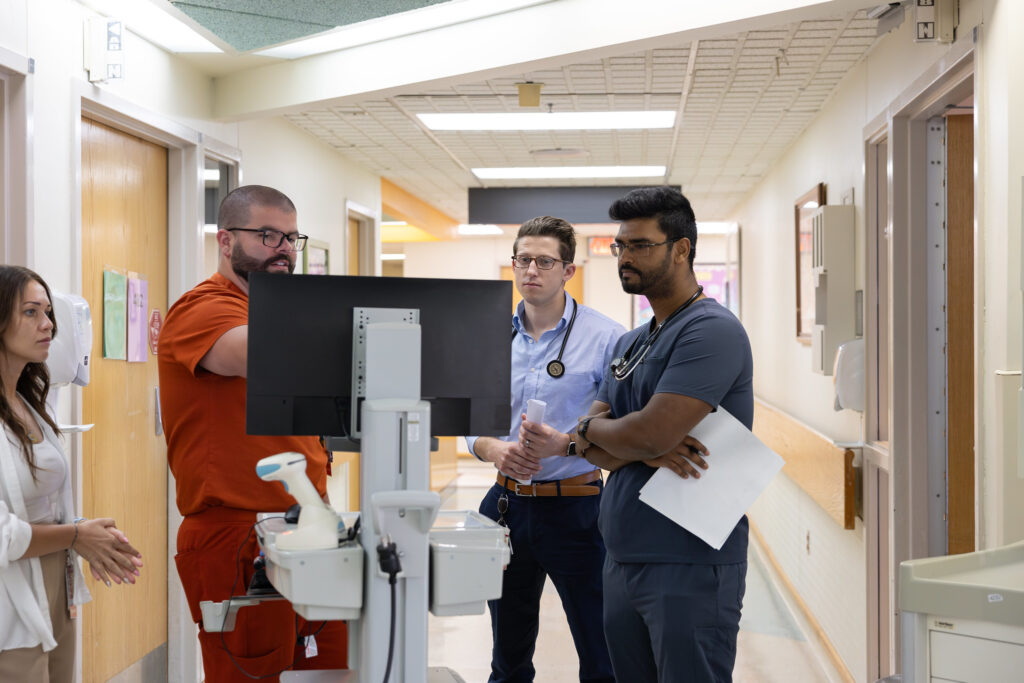 Richie stands in a hospital hallway, with colleagues, chaatting around a portable medical records system to discuss a patient. 