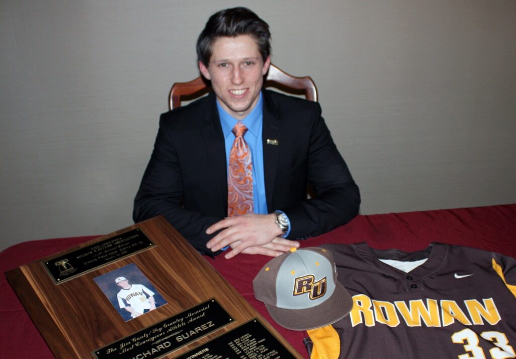 A college-aged Richie sits at a desk wearing a formal suit and tie, surrounded by Rowan swag such as his then-upcoming baseball jersey, a hat, and more. 